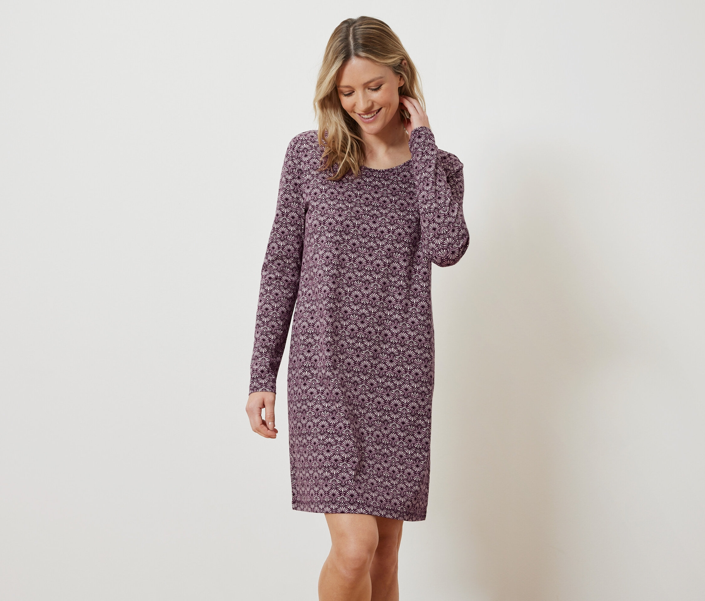 Une femme blonde souriante pose, portant une robe longue à motifs floraux.