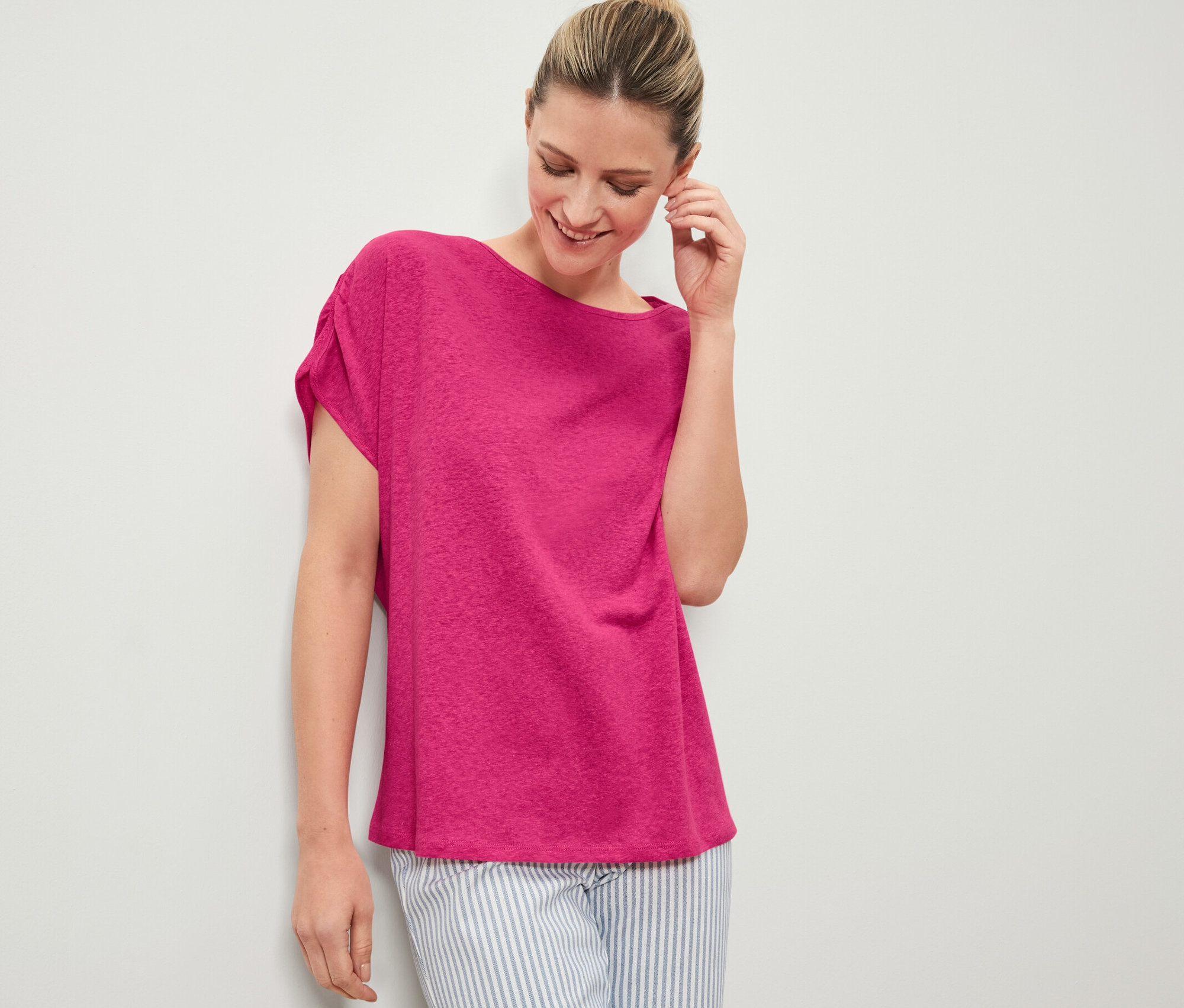 Femme souriant et touchant son oreille, elle porte un t-shirt rose en lin mélangé et un pantalon rayé.