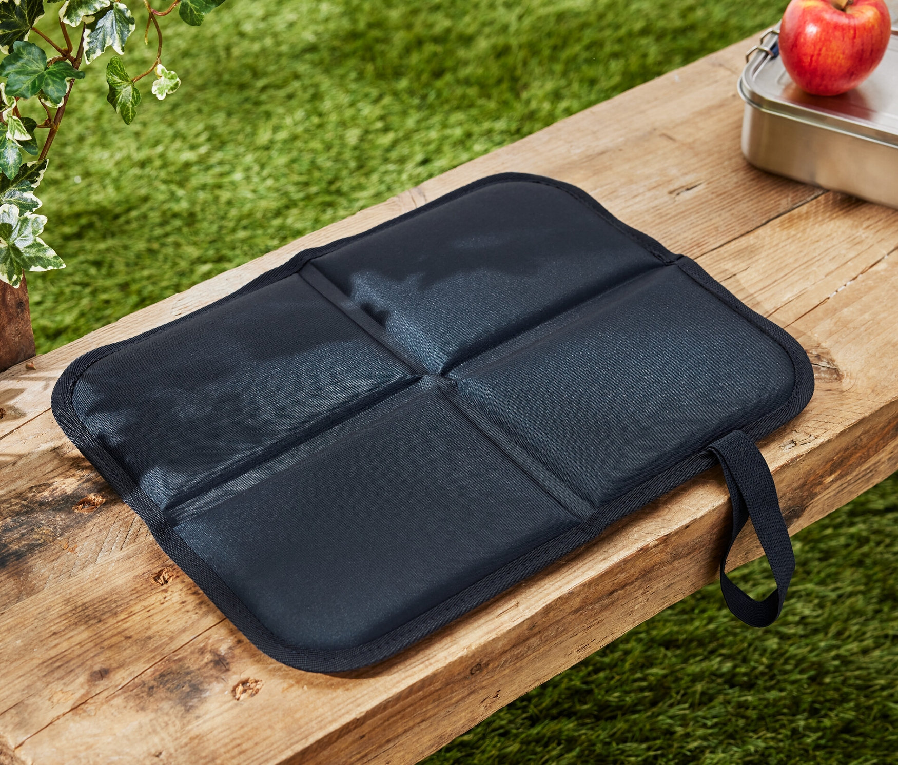Un coussin d’assise thermique pliable est posé sur un banc en bois à côté d’une pomme et d’une boîte à lunch.