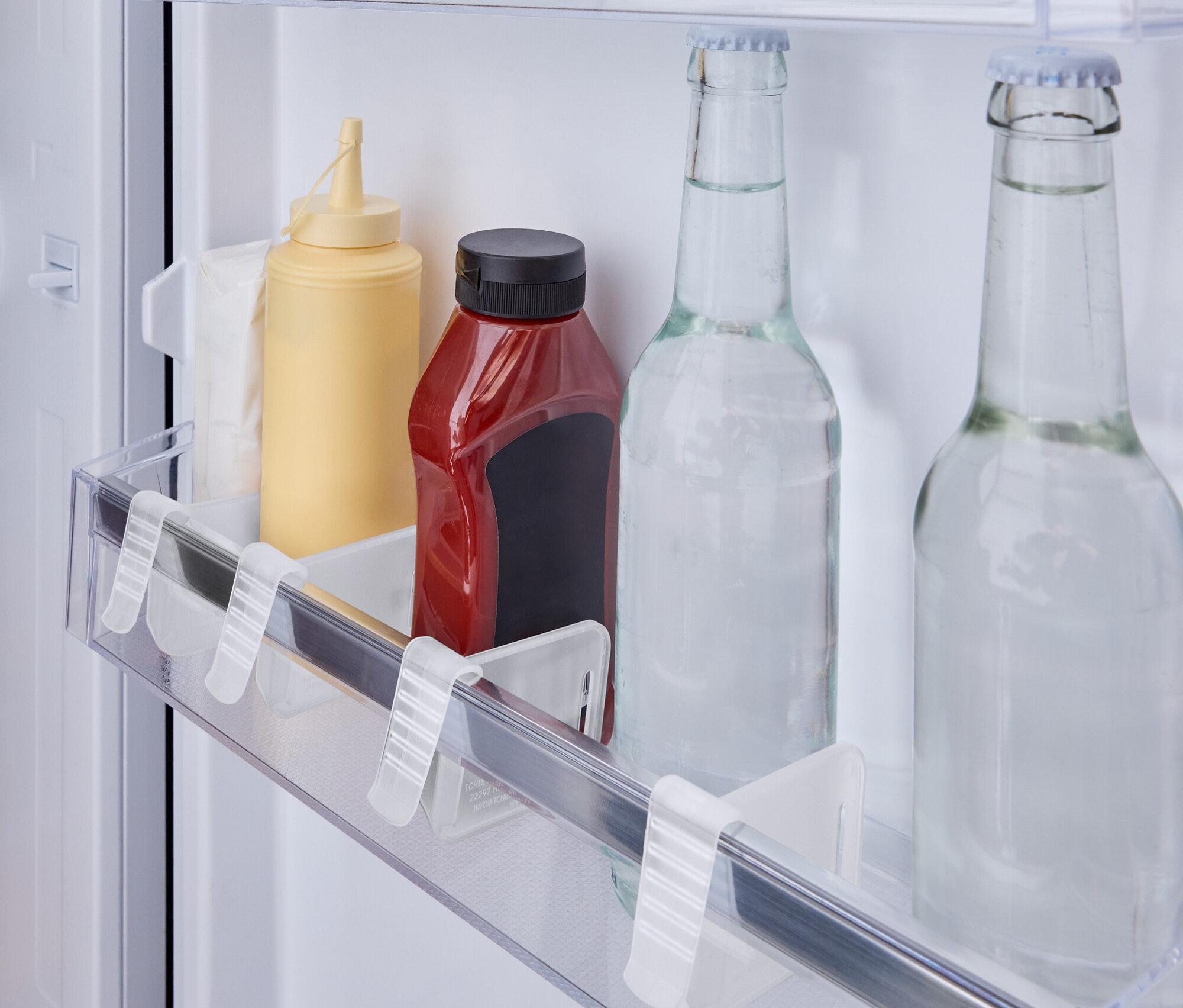 Dans le réfrigérateur, sur une étagère, se trouvent une bouteille de moutarde jaune, une bouteille de ketchup rouge et deux bouteilles d'eau en verre.