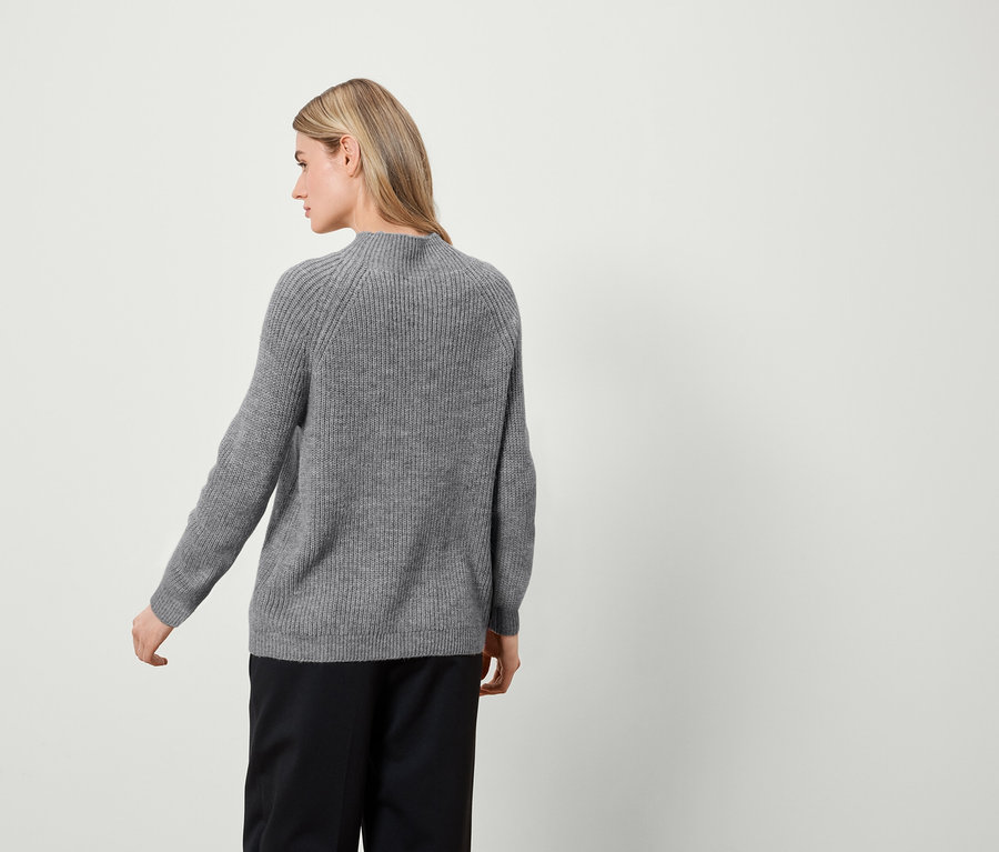 Une femme pose de dos, portant un pull gris et un pantalon noir.