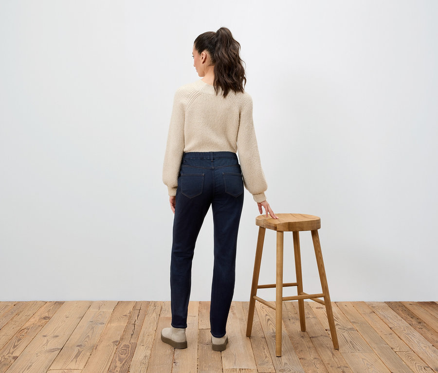 Une femme portant un pull beige et un jean bleu foncé se tient dos à la caméra, appuyée sur un tabouret en bois.