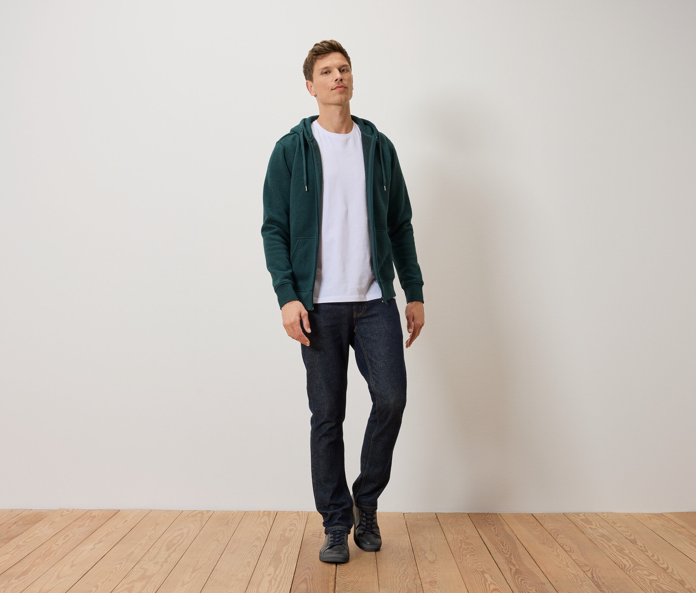 Un homme debout sur un plancher en bois, portant un jean bleu foncé, un t-shirt blanc et un sweat-shirt zippé vert.