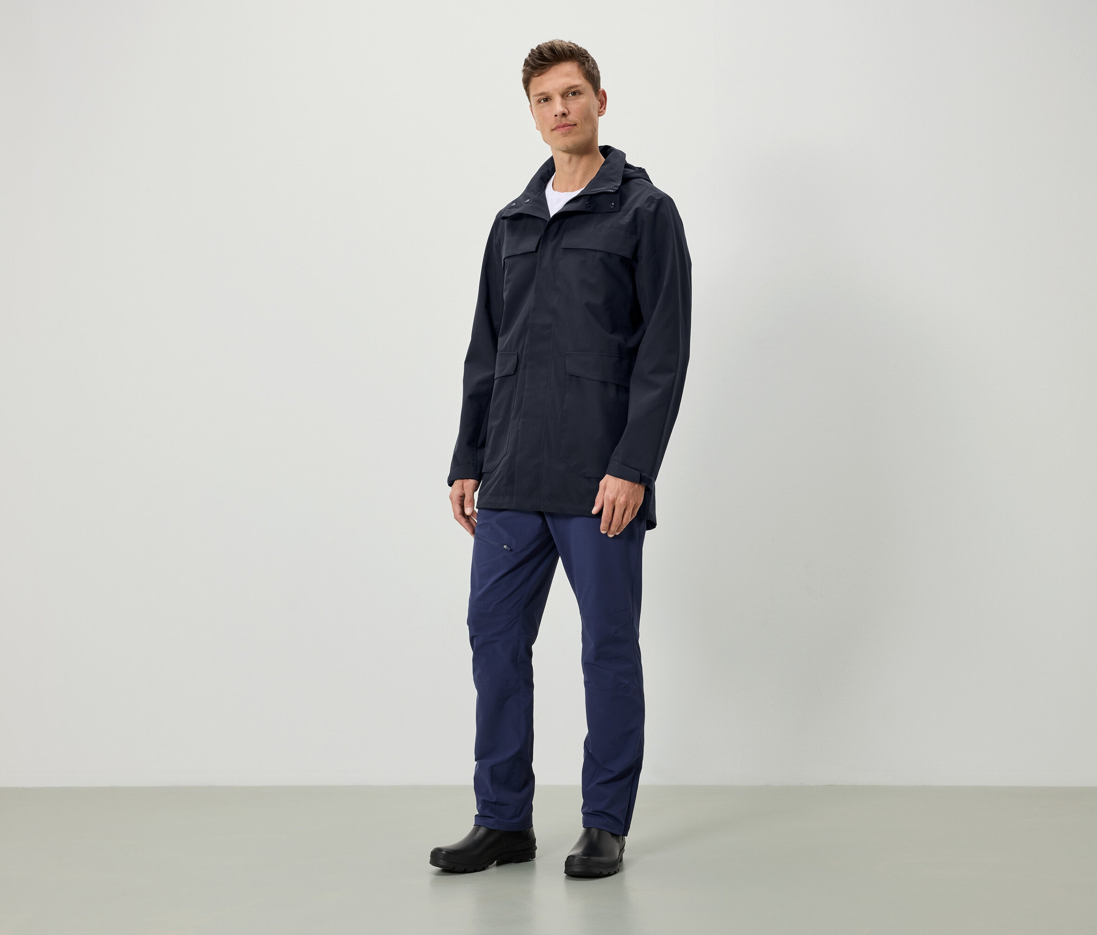 Un homme debout porte une veste bleu foncé à capuche, un pantalon bleu foncé et des chaussures noires.