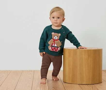 Un enfant se tient à côté d'un tabouret en bois, portant un pull vert avec un ours jouant du tambour et un pantalon marron.