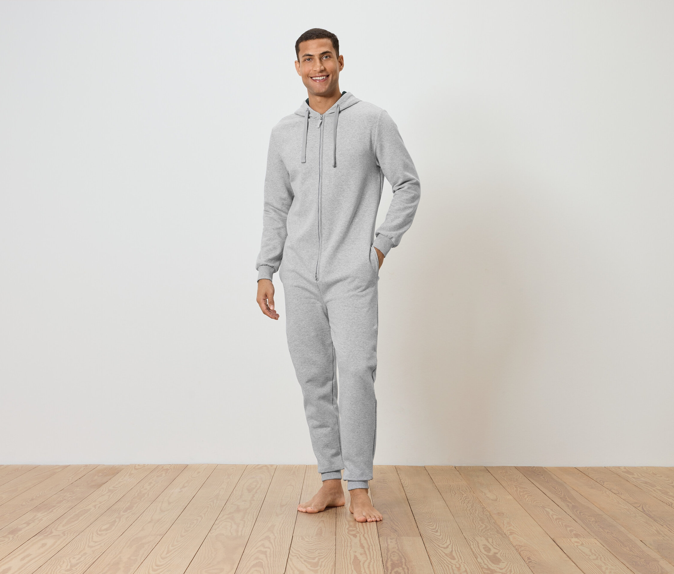Un homme debout pieds nus sur un plancher en bois, portant une combinaison grise à capuche, souriant.