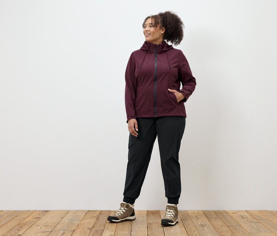 Une femme portant une veste zippée bordeaux, un pantalon noir et des chaussures marron se tient sur un plancher en bois.