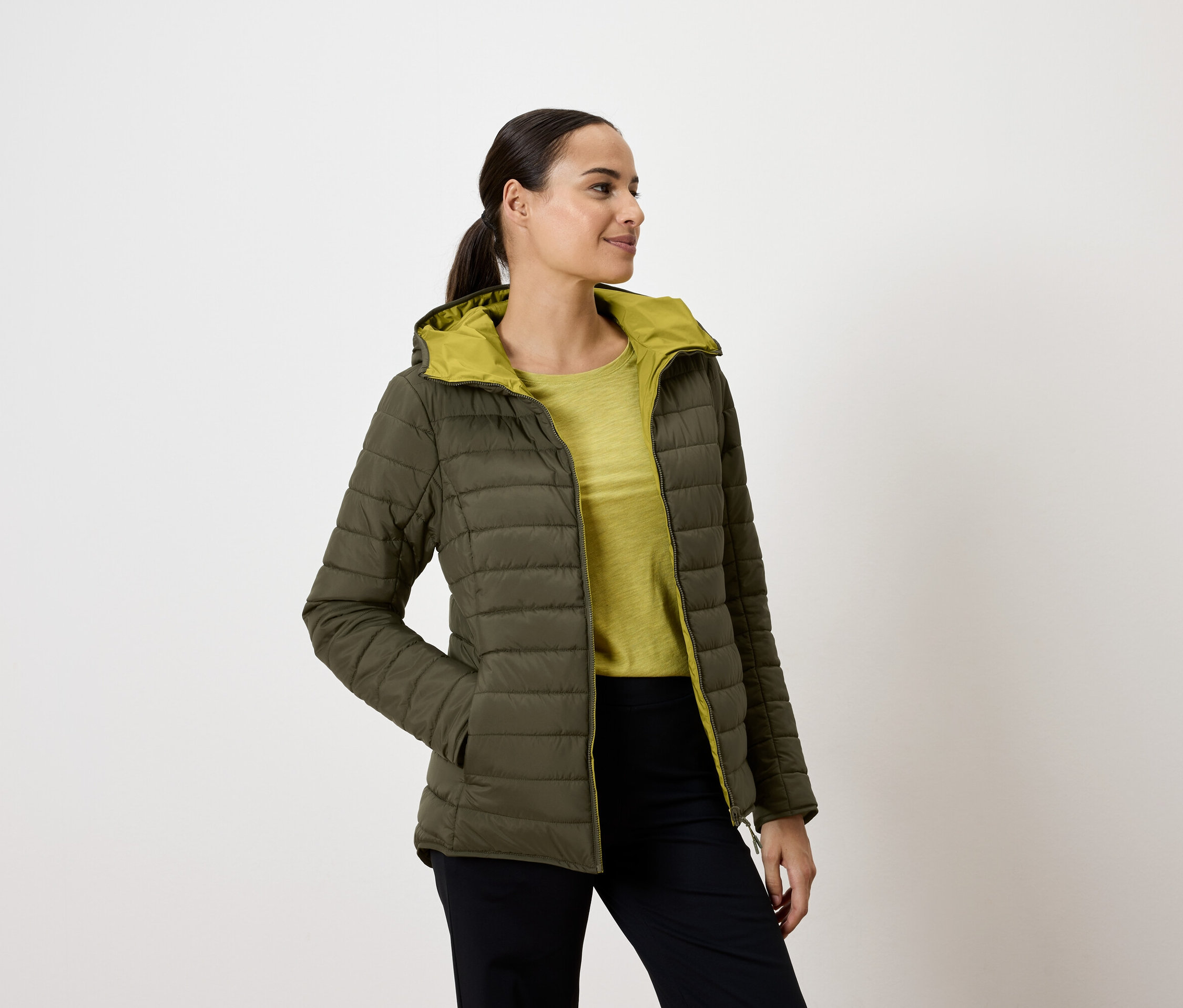 Une femme portant une veste matelassée vert olive à capuche, les mains dans les poches, regardant vers la droite.
