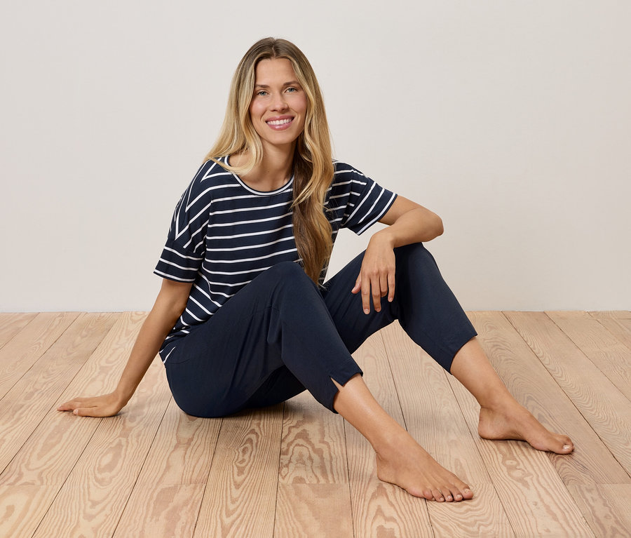 Une femme blonde est assise sur un plancher en bois, portant un t-shirt rayé bleu marine et un pantalon bleu foncé.