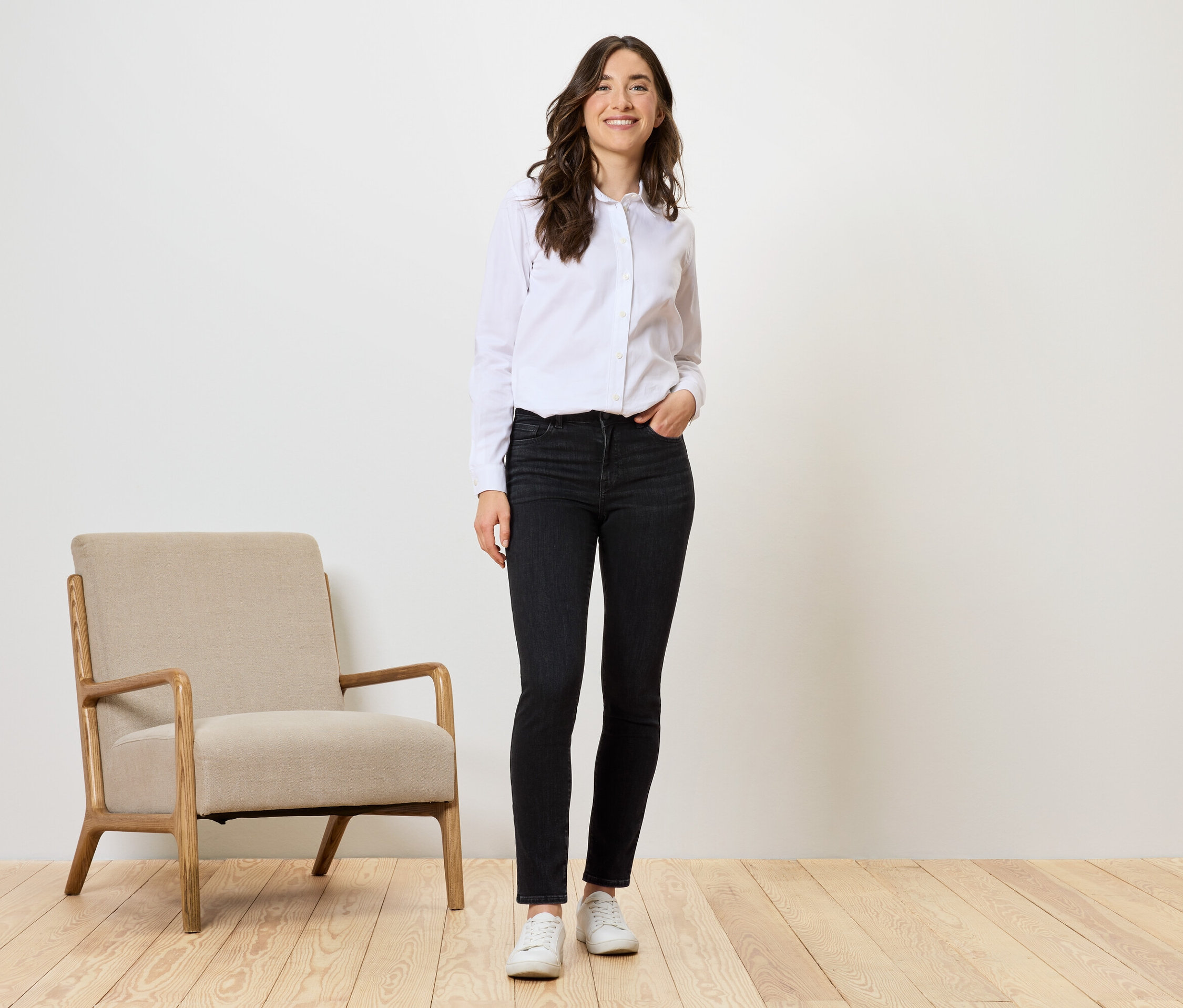 Une femme se tient debout en jean noir, chemise blanche et chaussures blanches à côté d'un fauteuil beige.