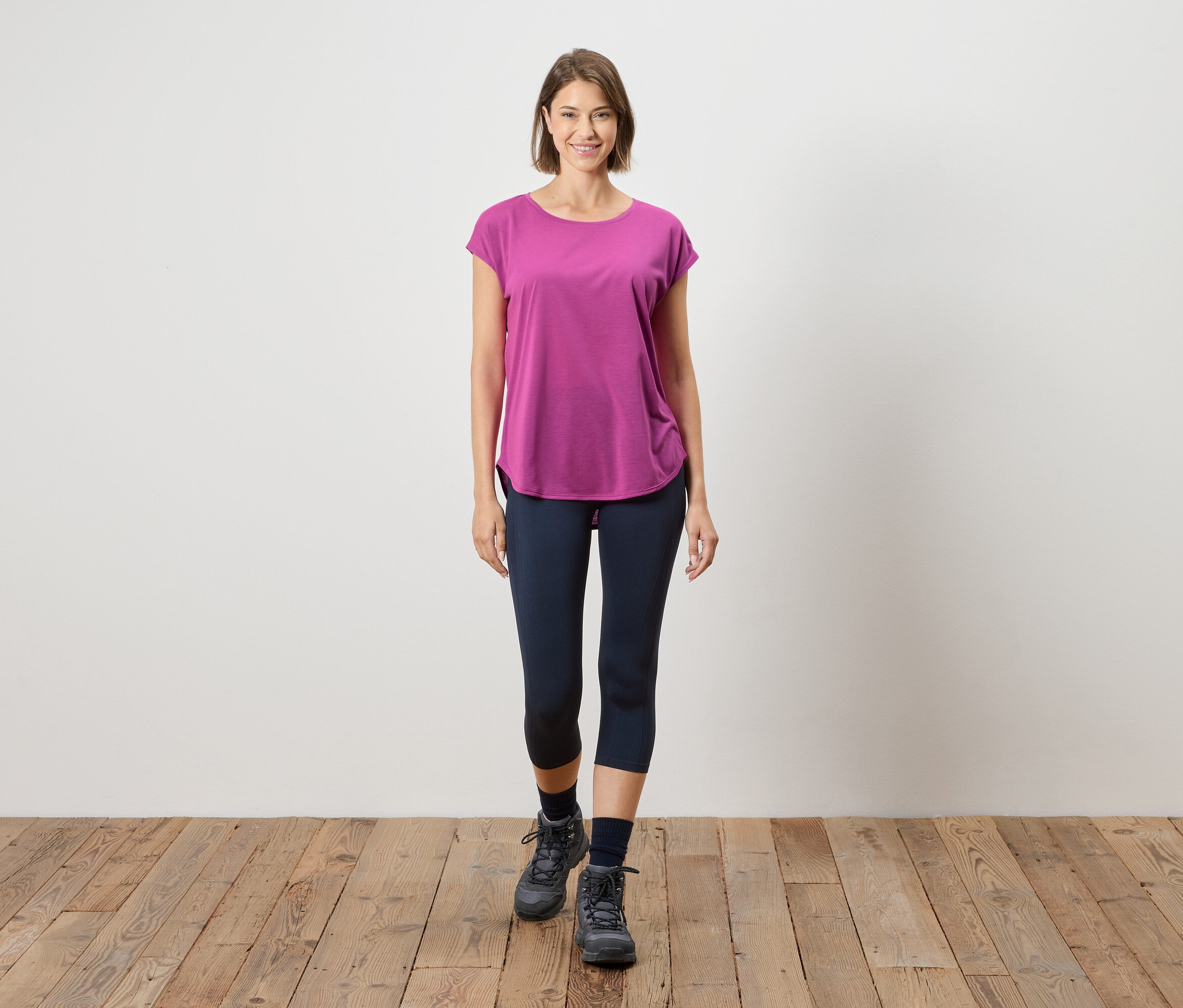 Une femme se tient sur un plancher en bois devant un mur blanc. Elle porte un t-shirt rose, des leggings bleus et des bottes grises.