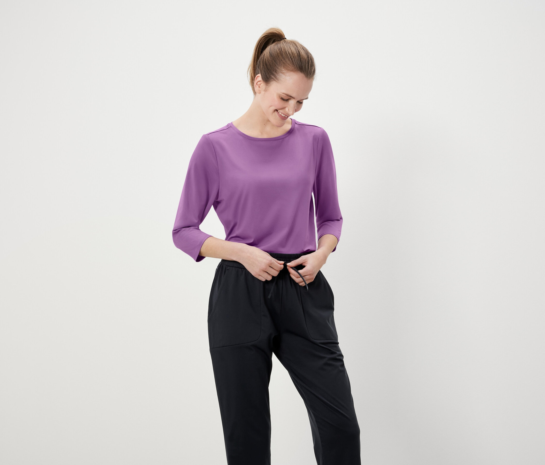 Une femme noue le cordon de serrage d'un pantalon noir. Elle porte un t-shirt violet à manches trois-quarts.