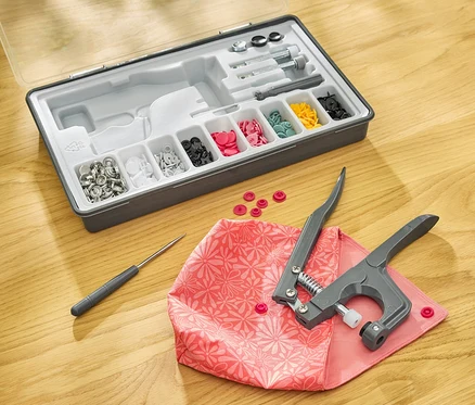 Un kit de boutons-pression sur une table en bois avec une pince à boutons-pression sur un tissu rose.