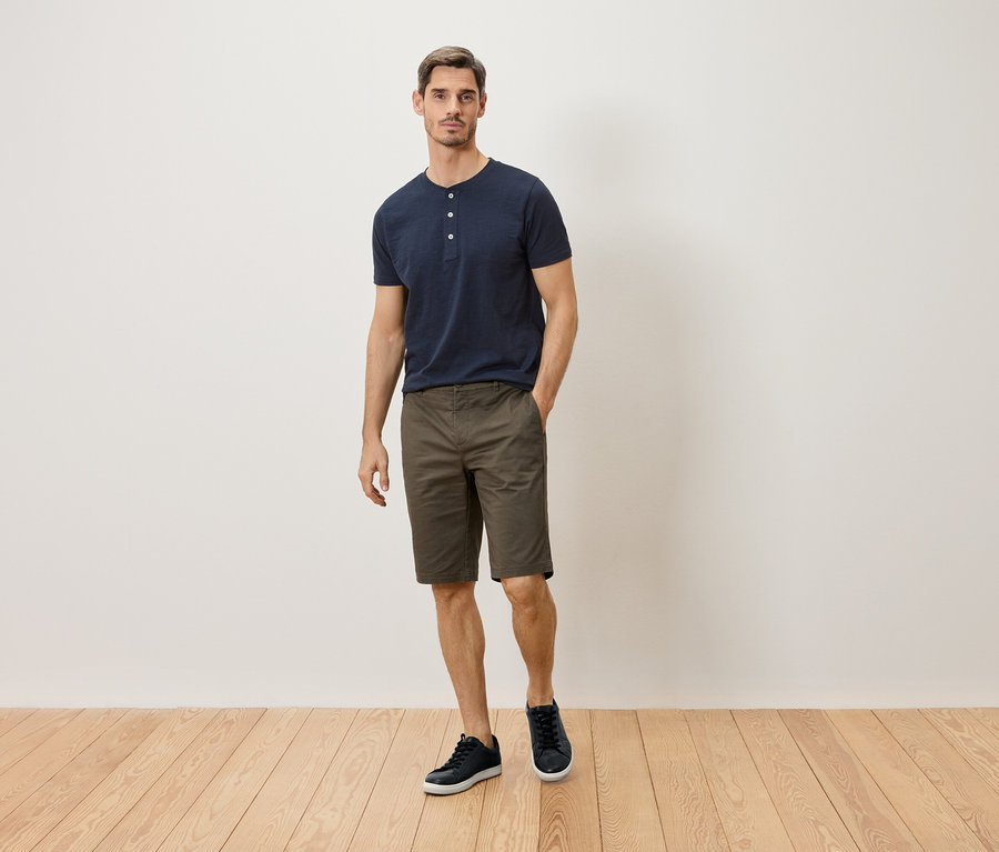 Un homme portant un t-shirt bleu marine boutonné, un short kaki et des baskets bleu marine se tient devant un mur blanc.