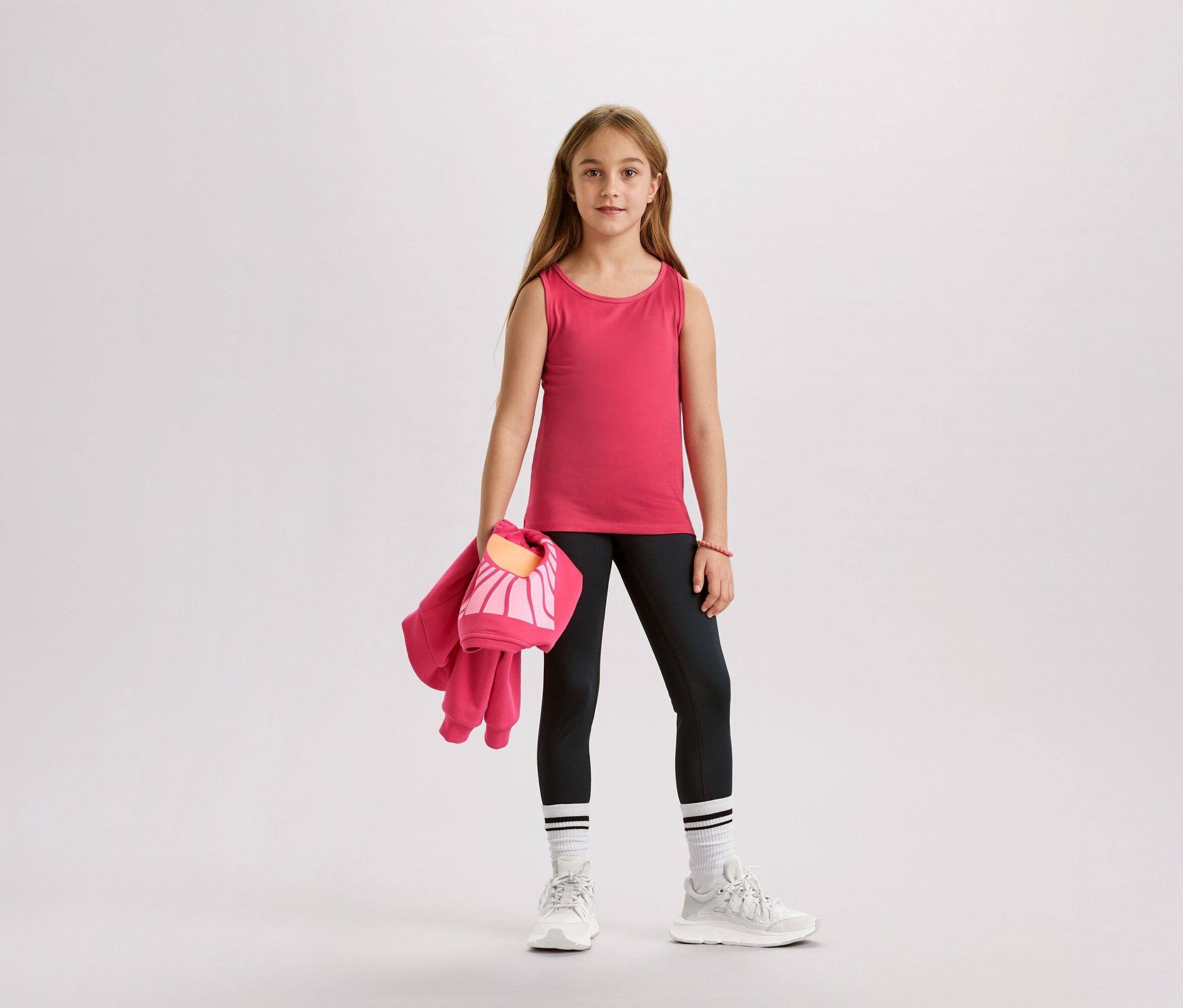 Fille portant un débardeur rose, un legging noir, des chaussettes blanches et des baskets, tenant une veste rose et un sac.