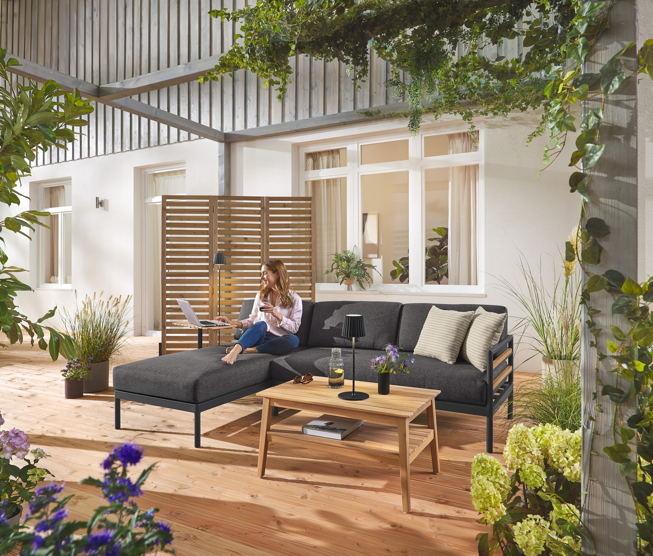 Une femme est assise sur le coin salon de jardin Premium « Elin » avec un paravent flexible en arrière-plan. On aperçoit également un lampadaire d'extérieur à LED sans fil noir et une lampe de table ainsi qu'une table basse de jardin.