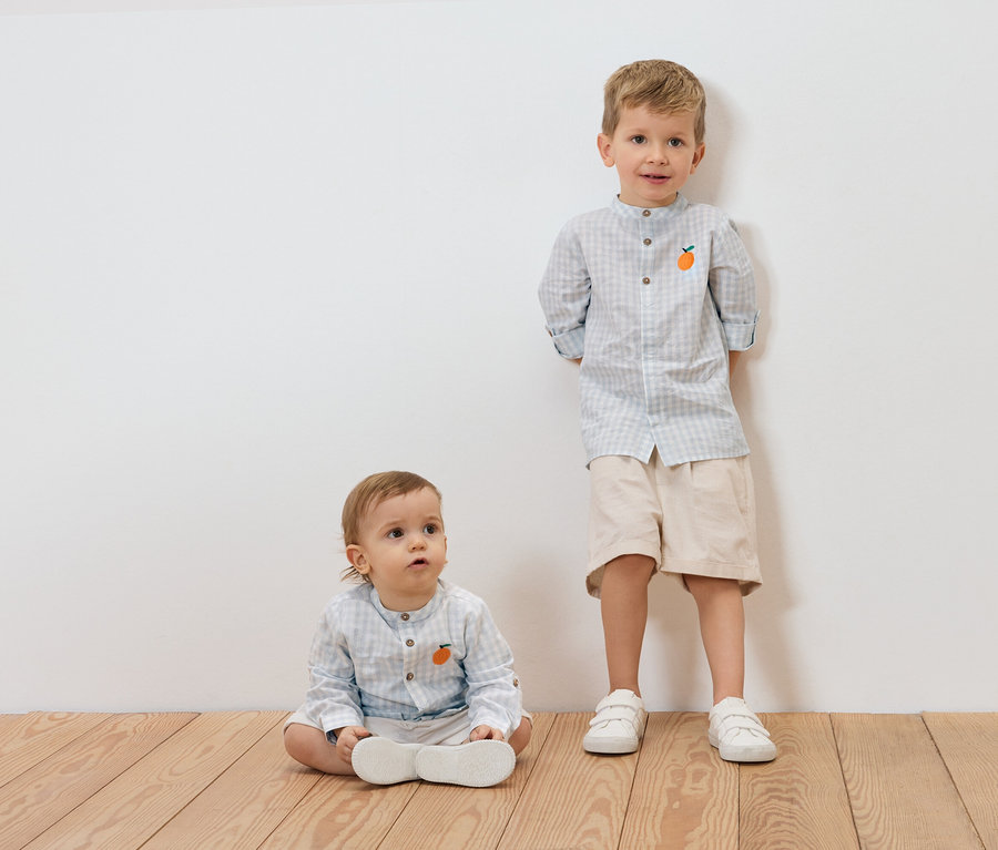 Un jeune enfant est assis sur un plancher en bois, à côté de lui se tient un garçon plus âgé, tous deux portant des chemises à carreaux bleu clair avec une application orange.