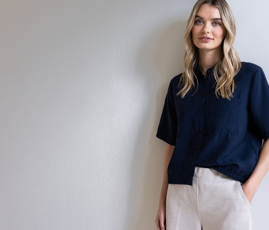 Une femme blonde portant une chemise bleu marine et un pantalon beige pose devant un mur gris clair.