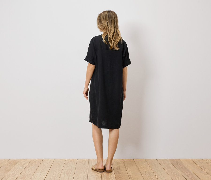 Vue de dos d'une femme aux cheveux blonds et ondulés portant une robe t-shirt noire jusqu'aux genoux et des sandales marron.