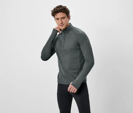 Un homme aux cheveux bouclés porte un T-shirt gris à manches longues avec fermeture éclair et un legging noir sur fond blanc.
