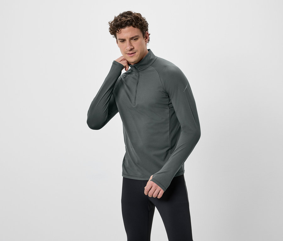 Un homme aux cheveux bouclés porte un T-shirt gris à manches longues avec fermeture éclair et un legging noir sur fond blanc.