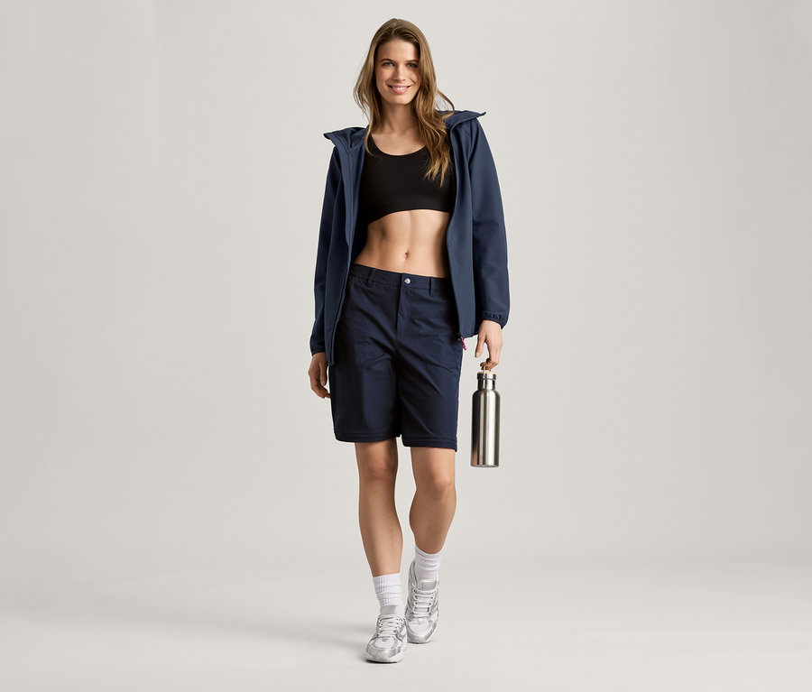 Une femme portant une veste en softshell bleu marine, un short et un soutien-gorge de sport tient une bouteille argentée.