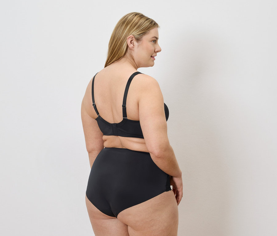 Une femme blonde, vue de dos, porte des sous-vêtements noirs sur un fond blanc.