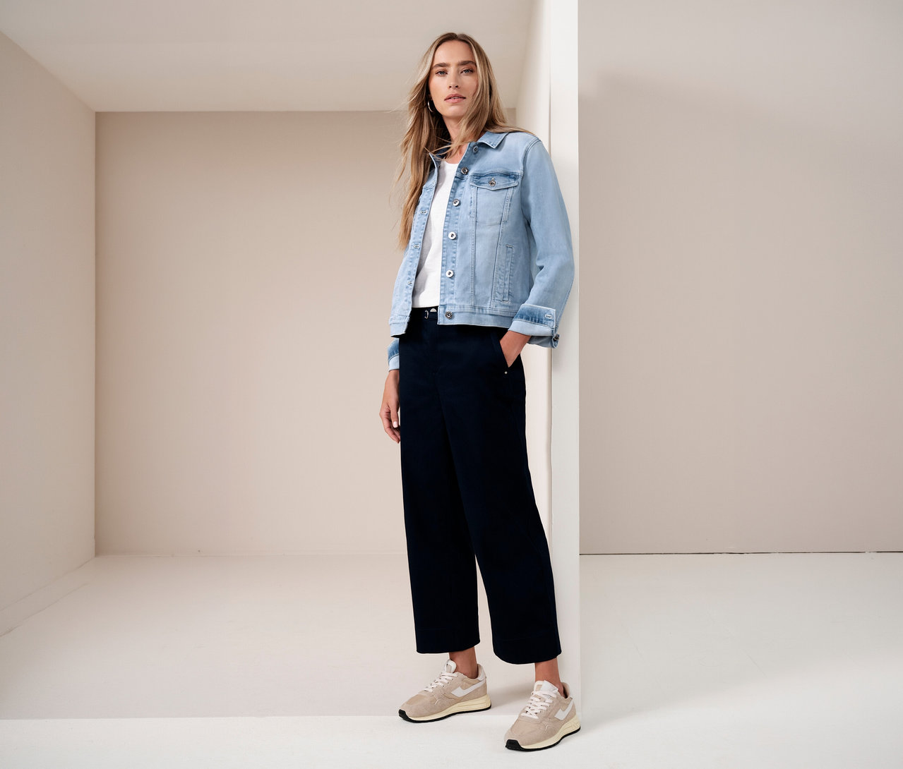 Une femme en veste en jean, t-shirt blanc et pantalon foncé, une main dans la poche, pose dans un coin d'une pièce lumineuse.