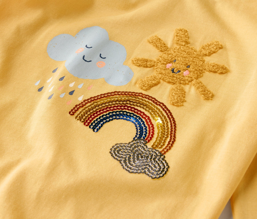 Sur un tissu jaune sont brodés un nuage avec des gouttes de pluie, un soleil et un arc-en-ciel.