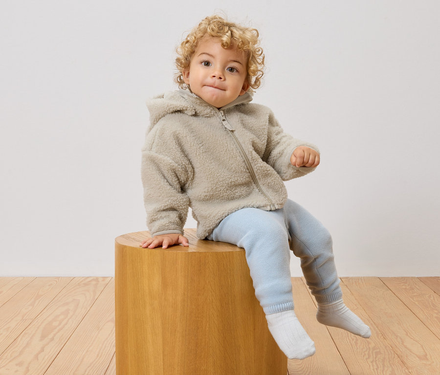 Un jeune enfant aux cheveux bouclés est assis sur un tabouret en bois, portant une veste polaire à capuche, un pantalon bleu clair et des chaussettes blanches.