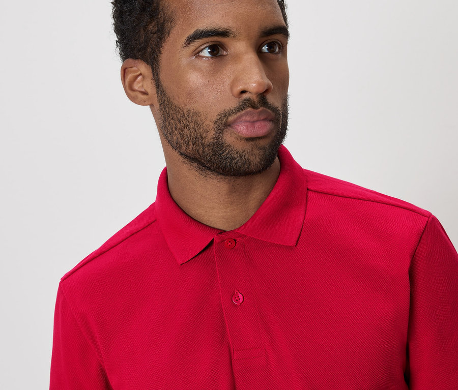 Un homme aux cheveux foncés et à la barbe porte un polo rouge sur fond blanc.
