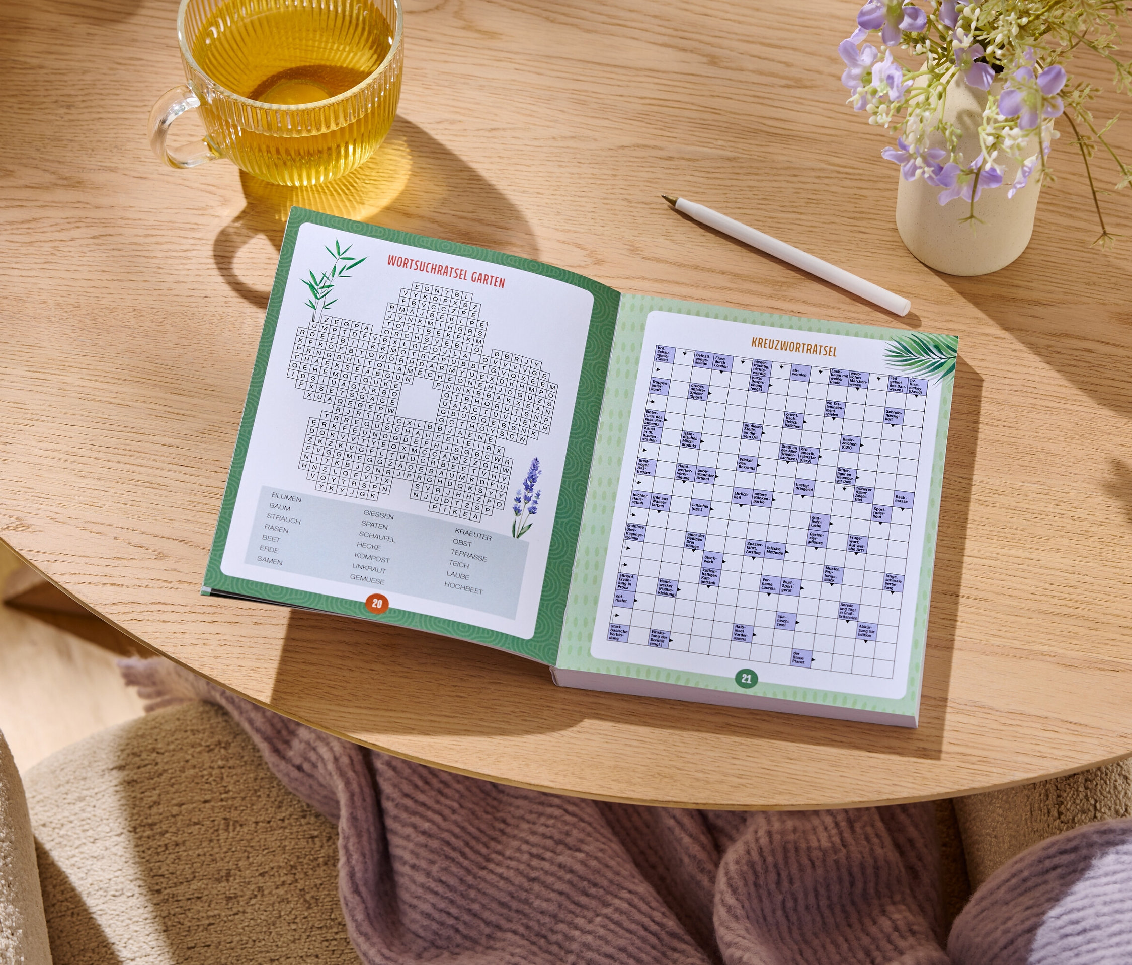 Sur une table en bois se trouve un livre de jeux ouvert à côté d'un verre de thé, d'un stylo et d'un petit vase de fleurs.