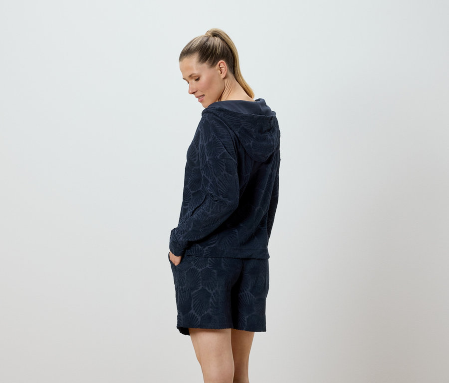 Femme posant en sweat à capuche et short à motif coquillage, les mains dans les poches.