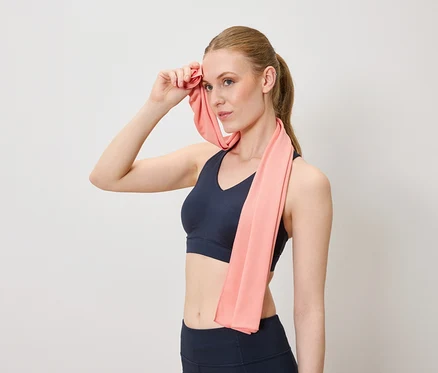 Une femme s'éponge le front avec une serviette rose, portant des vêtements de sport sur un fond neutre.