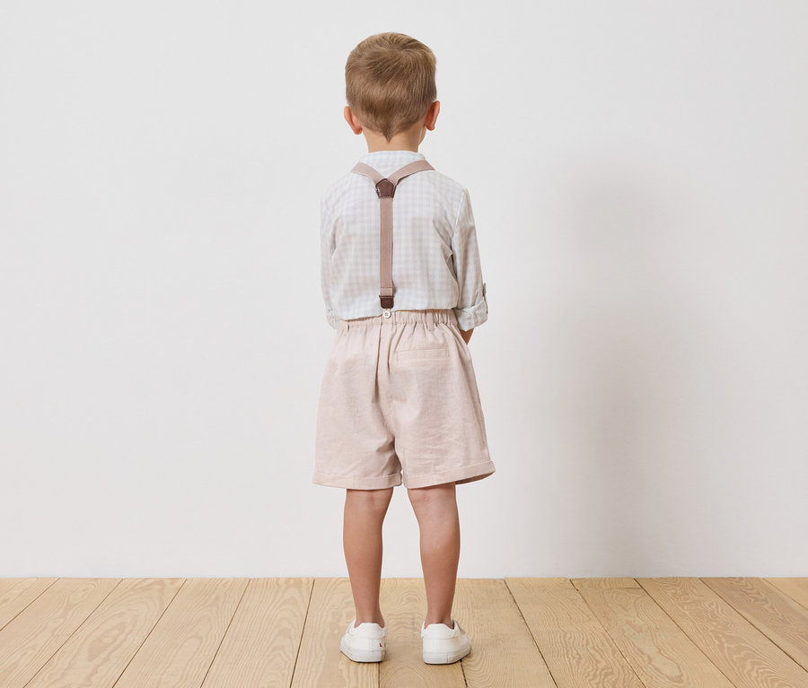 Un enfant se tient dos à la caméra, portant une chemise à carreaux, un short et des bretelles.