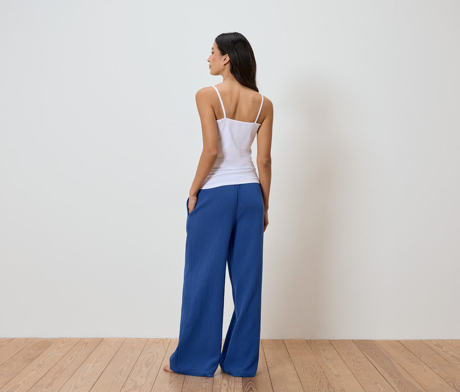 Femme vue de dos portant un débardeur blanc et un pantalon bleu devant un mur blanc.