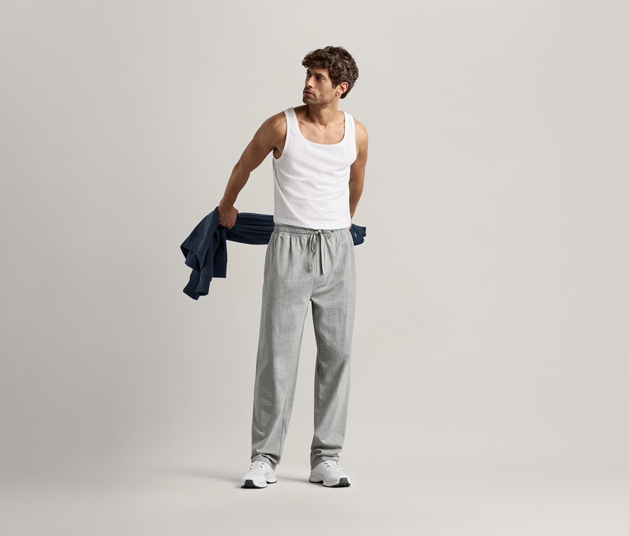 Un homme en débardeur et pantalon relax gris tenant une chemise bleue sur le bras.