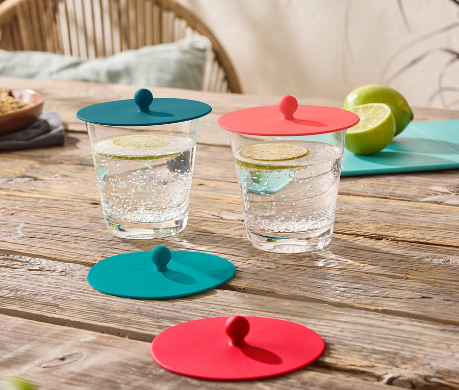 Deux verres de boissons sont posés sur une table en bois, chacun fermé par un couvercle en silicone, un turquoise et un rouge.