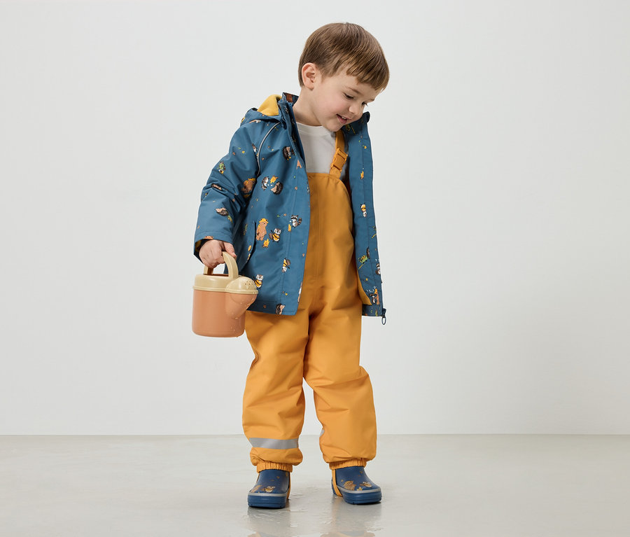 Un garçon aux cheveux bruns porte une veste bleue à motifs d'animaux, une salopette jaune et des bottes bleues, et tient un arrosoir.