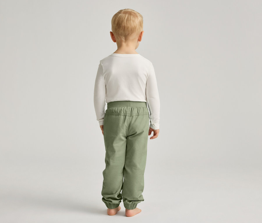 Un enfant blond est vu de dos, portant un t-shirt blanc à manches longues et un pantalon vert.