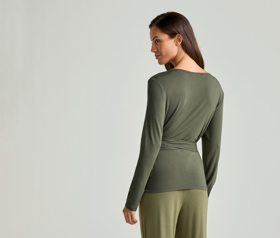 Une femme aux longs cheveux bruns porte un haut à manches longues vert olive et un pantalon, regardant par-dessus son épaule.