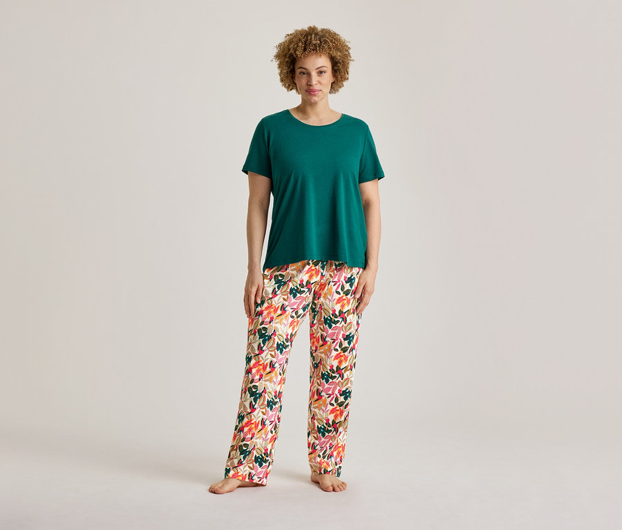 Une femme aux cheveux bouclés est debout, vêtue d'un t-shirt vert et d'un pantalon de pyjama à motifs floraux d'un ensemble de pyjama, pieds nus.