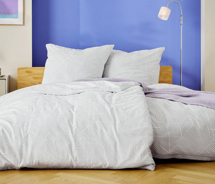 Un lit avec des draps blancs à motifs géométriques et deux oreillers se dresse devant un mur bleu.