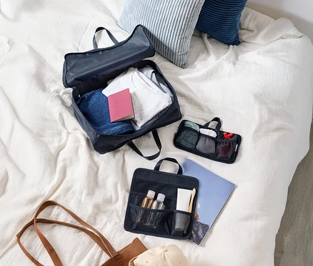 Vue de dessus d'une valise avec des vêtements, deux coussins, une trousse de toilette, un sac à main et une casquette sur un lit.