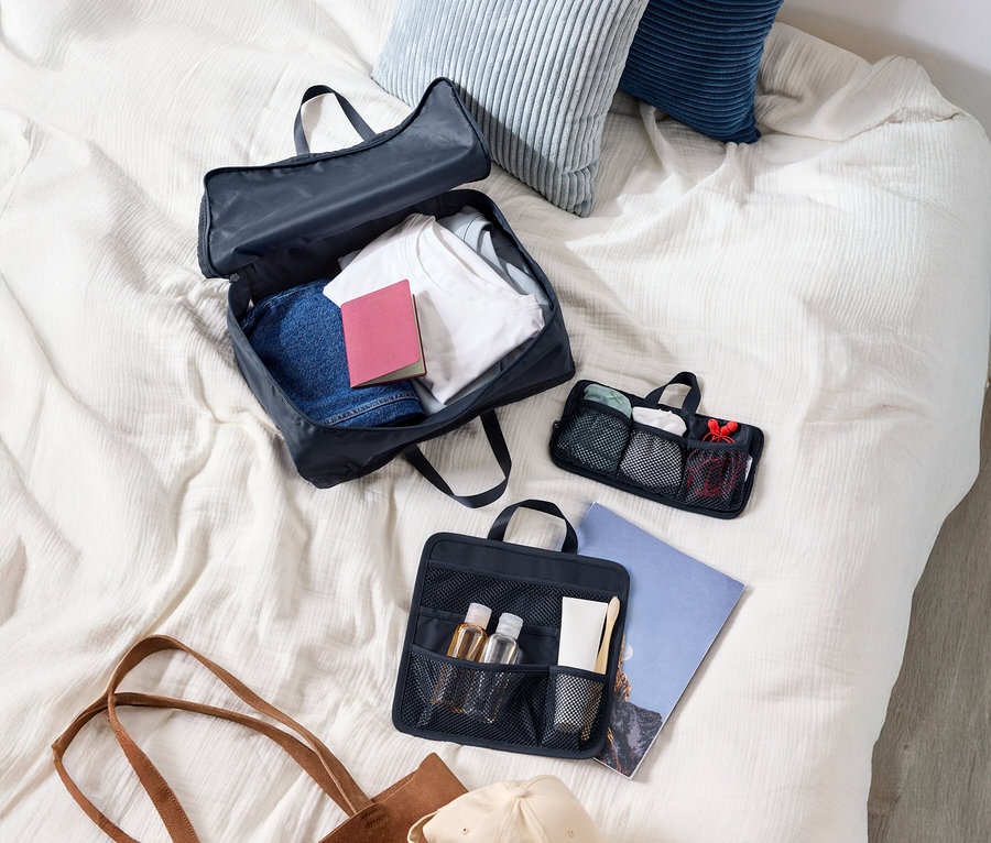 Vue de dessus d'une valise avec des vêtements, deux coussins, une trousse de toilette, un sac à main et une casquette sur un lit.