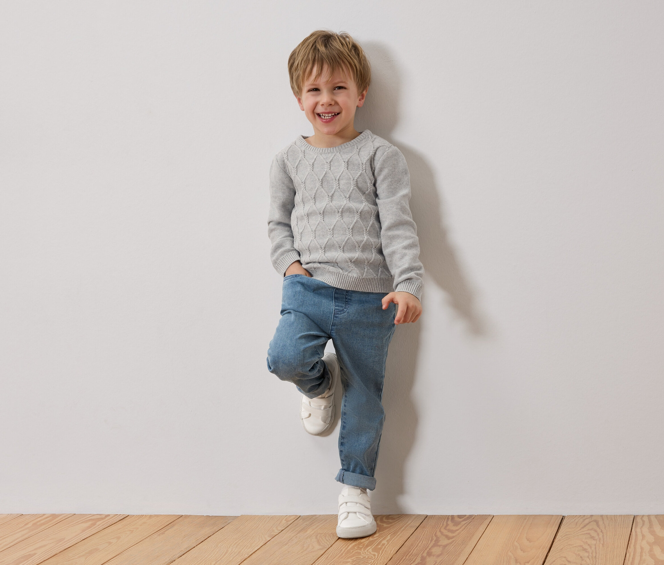 Un garçon blond souriant est appuyé contre un mur blanc. Il porte un pull gris, un jean bleu et des chaussures blanches.