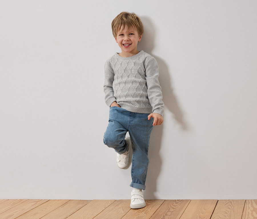 Un garçon blond souriant est appuyé contre un mur blanc. Il porte un pull gris, un jean bleu et des chaussures blanches.