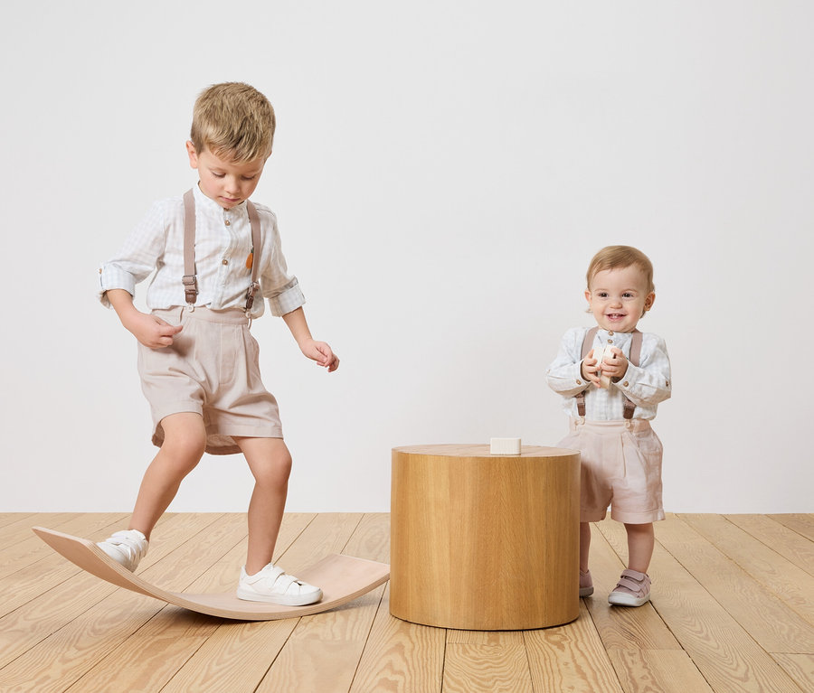 Deux enfants, un garçon en équilibre sur une planche en bois, un bébé debout à côté d'une table en bois tenant un bloc de jouet.