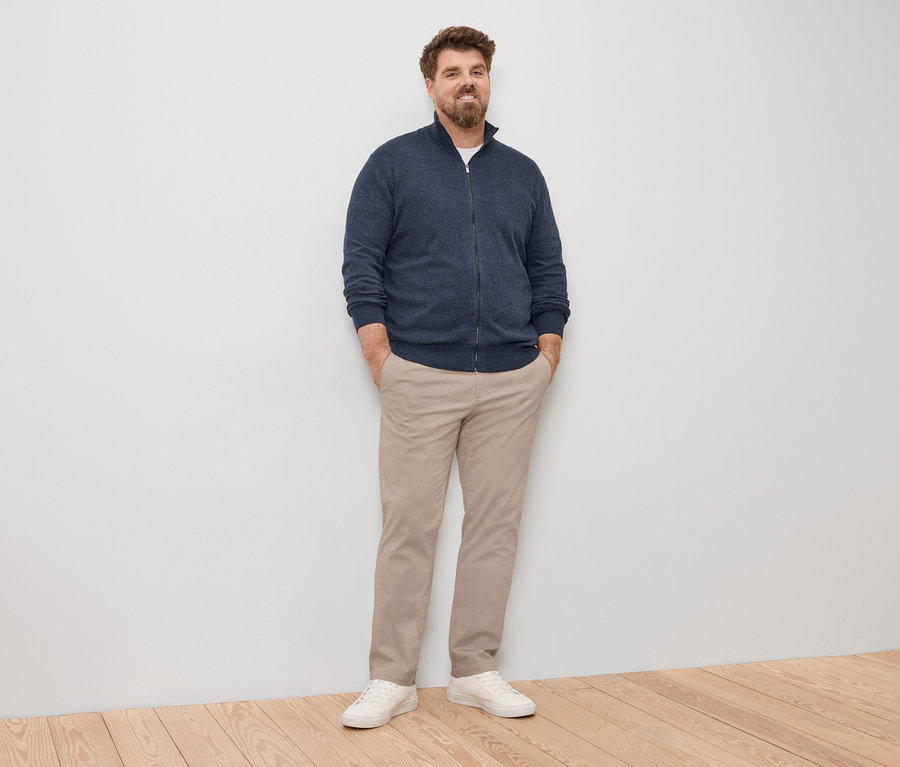 Un homme se tient debout, les mains dans les poches, portant un cardigan en maille bleu marine, un pantalon clair et des baskets blanches.
