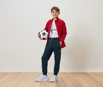 Un garçon tient un ballon de foot dans ses mains. Il porte une veste rouge, un pantalon et des baskets blanches.